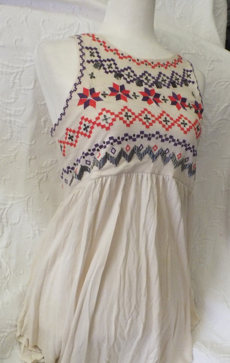 May include: A sleeveless, cream-colored top with a geometric pattern embroidered in red, purple, and black. The top has a gathered neckline and a loose, flowing fit.