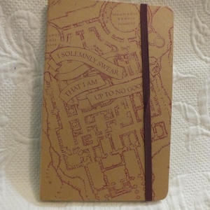 May include: A brown notebook with a red and white map of Hogwarts on the cover. The map includes the text "I solemnly swear that I am up to no good". The notebook has a brown elastic band.