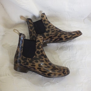 May include: A pair of ankle-high rain boots with a leopard print pattern. The boots have a tan base color with dark brown spots, black elastic side panels, and small pull tabs. The boots are made of a glossy material.