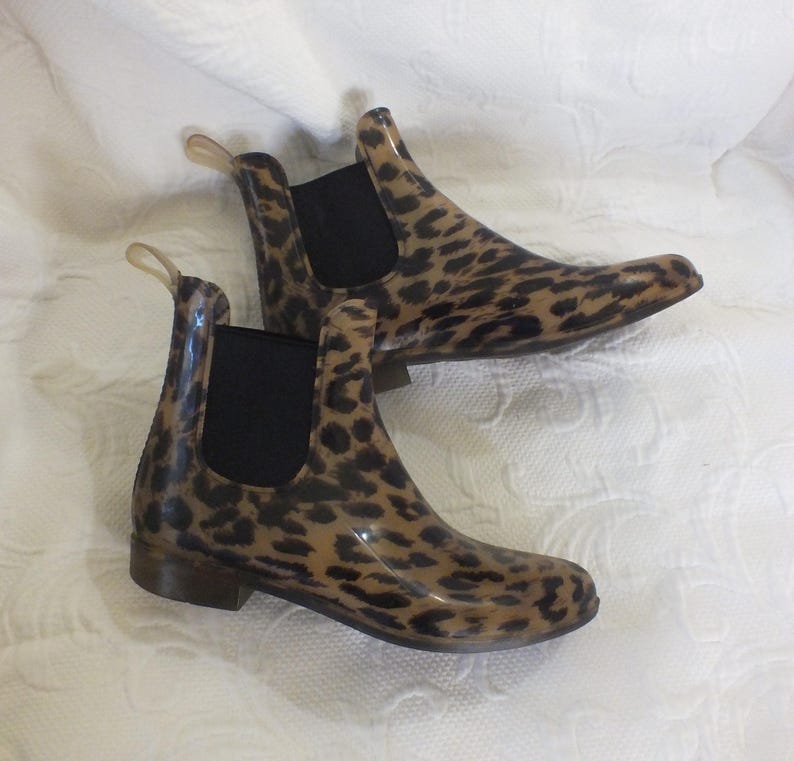 May include: A pair of ankle-high rain boots with a leopard print pattern. The boots have a tan base color with dark brown spots, black elastic side panels, and small pull tabs. The boots are made of a glossy material.