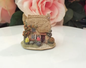 Może przedstawiać: Miniaturowa figurka wiejskiego domku z dachem z trzciny, brązowymi drzwiami, dwoma oknami i małym ogrodem z kwiatami. Domek jest wykonany z jasnobrązowego materiału i stoi na białym tle.