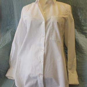 May include: A white button-down shirt with long sleeves, a collar, and a chest pocket. The shirt is displayed on a mannequin against a blue-green background. The shirt has a classic, tailored design.