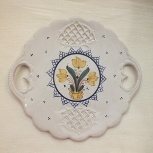 Op de afbeelding: Wit keramisch serveerplateau met geschulpte randen en twee handvatten. Het midden heeft een bloemenontwerp met gele tulpen in een gele vaas, omgeven door een blauw-witte rand. Decoratieve roosterpatronen.