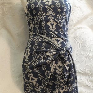 May include: Sleeveless navy blue dress with a white damask pattern. The dress features a knotted detail at the waist. The dress is displayed on a mannequin.