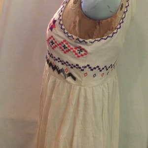 May include: A white sleeveless dress with a colorful geometric pattern embroidered on the bodice. The dress has a gathered skirt and a relaxed fit.