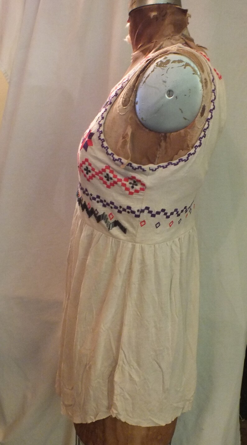 May include: A white sleeveless dress with a colorful geometric pattern embroidered on the bodice. The dress has a gathered skirt and a relaxed fit.