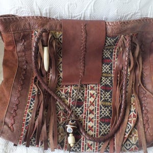 May include: Brown leather tote bag with a woven fabric center panel featuring geometric patterns in red, black, and cream. The bag has leather fringe, a braided handle, and a flap closure.