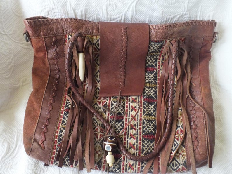 May include: Brown leather tote bag with a woven fabric center panel featuring geometric patterns in red, black, and cream. The bag has leather fringe, a braided handle, and a flap closure.