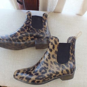 May include: A pair of ankle-high rain boots with a leopard print pattern. The boots have a brown and black spotted design, a black elastic panel on the sides, and a small heel. The boots are made of a glossy material.