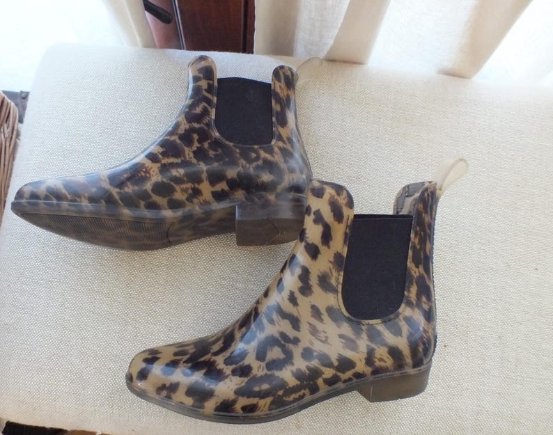 May include: A pair of ankle-high rain boots with a leopard print pattern. The boots have a brown and black spotted design, a black elastic panel on the sides, and a small heel. The boots are made of a glossy material.