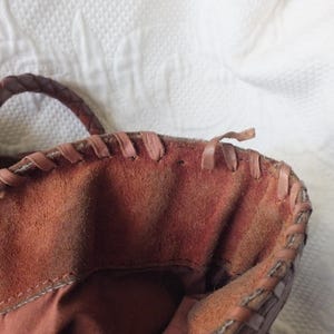 May include: Close-up of a brown leather tote bag. The bag features a braided handle and stitched detailing along the top edge. The interior is a darker shade of brown, and the bag appears to be made of suede or a similar material.