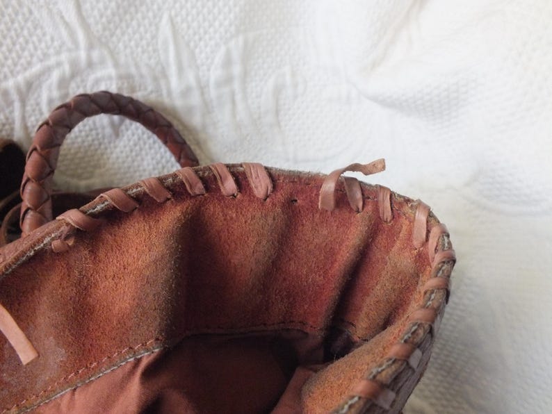May include: Close-up of a brown leather tote bag. The bag features a braided handle and stitched detailing along the top edge. The interior is a darker shade of brown, and the bag appears to be made of suede or a similar material.