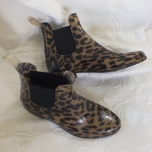 May include: A pair of ankle-high rain boots with a leopard print pattern. The boots have a brown and black spotted design on a clear, glossy material. They feature black elastic side panels and a clear pull tab at the back.