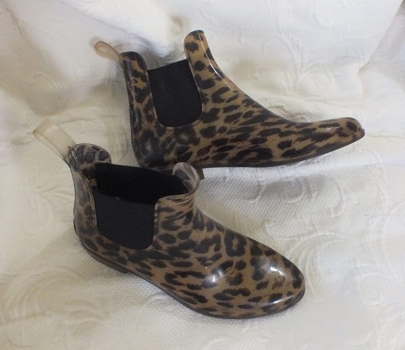 May include: A pair of ankle-high rain boots with a leopard print pattern. The boots have a brown and black spotted design on a clear, glossy material. They feature black elastic side panels and a clear pull tab at the back.