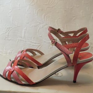 May include: A pair of strappy, open-toe sandals in shades of pink and coral. The heels are a vibrant coral color, and the straps feature a snakeskin pattern. The sandals have a gold buckle closure and are displayed on a white box.