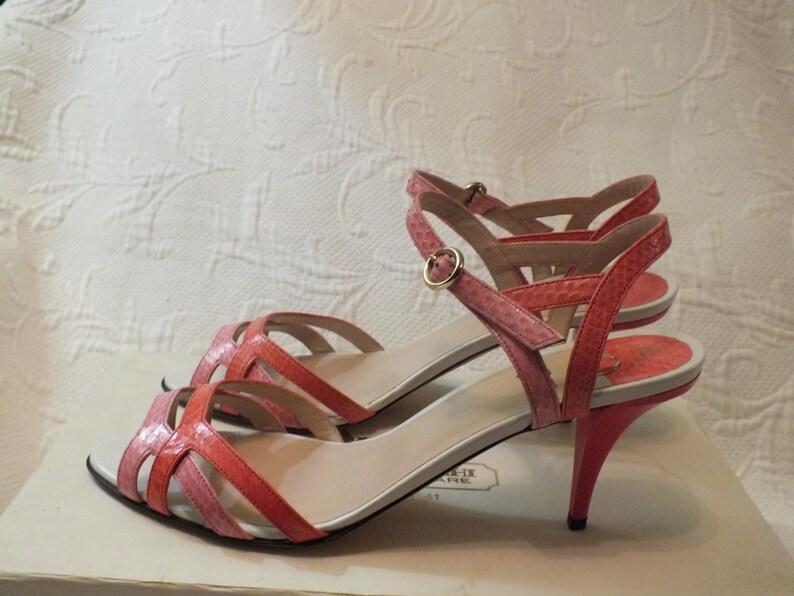 May include: A pair of strappy, open-toe sandals in shades of pink and coral. The heels are a vibrant coral color, and the straps feature a snakeskin pattern. The sandals have a gold buckle closure and are displayed on a white box.