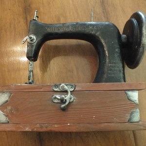 May include: A vintage black sewing machine with a wooden base and a metal latch. The machine is labeled "Willcox & Gibbs M. Co."