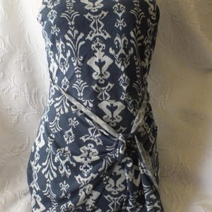 May include: Sleeveless navy blue dress with a white damask pattern. The dress features a knotted waist detail and a scoop neckline. The fabric has a soft, flowing drape, and the overall style is elegant and versatile.