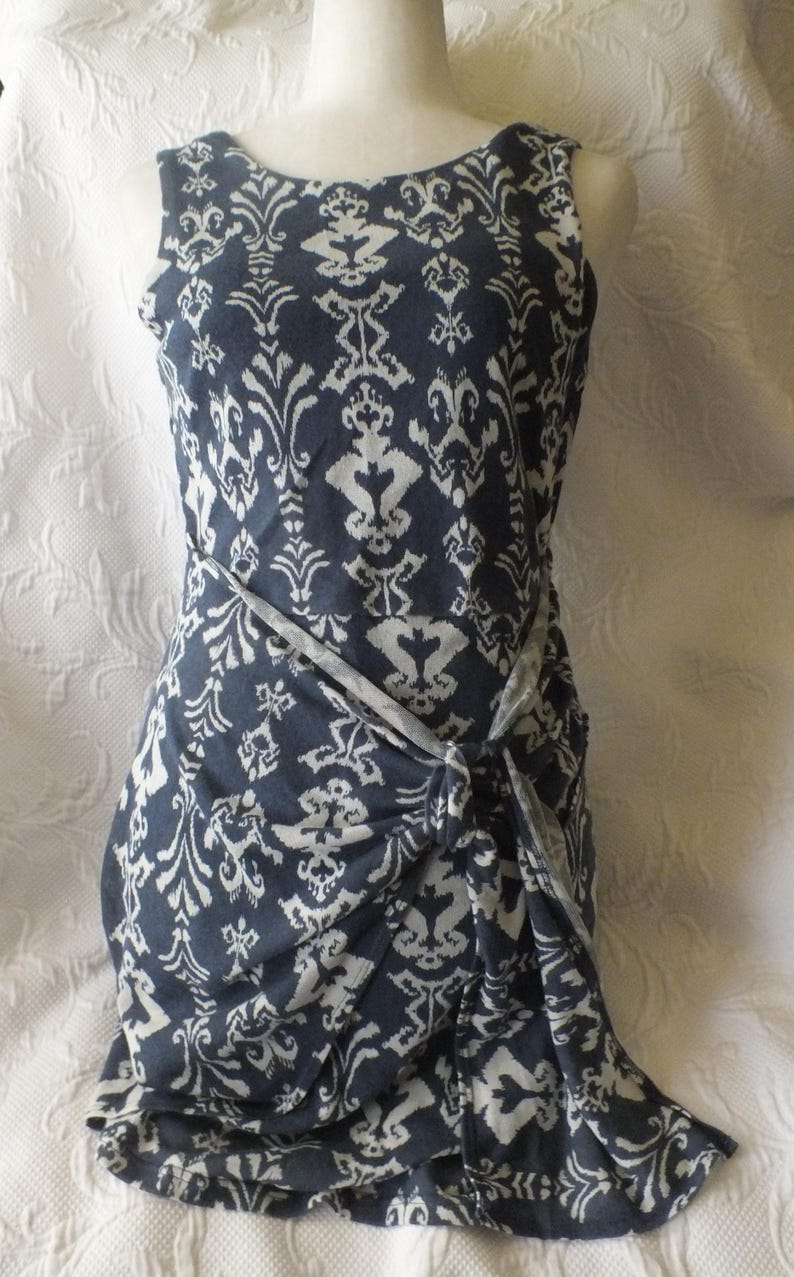 May include: Sleeveless navy blue dress with a white damask pattern. The dress features a knotted waist detail and a scoop neckline. The fabric has a soft, flowing drape, and the overall style is elegant and versatile.