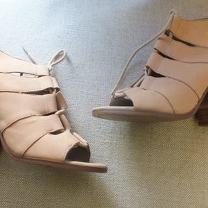 Könnte beinhalten: Ein Paar beige Sandalen mit offenem Zehenbereich und Blockabsatz. Die Sandalen haben mehrere Riemen über Fuß und Knöchel mit Schnürverschluss. Die Absätze sind etwa 7,6 cm hoch.