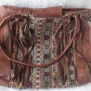 May include: Brown leather shoulder bag with a woven fabric panel featuring geometric patterns in red, black, and gold. The bag has long leather fringe and a braided handle. The bag is a boho style.