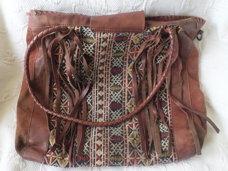 May include: Brown leather shoulder bag with a woven fabric panel featuring geometric patterns in red, black, and gold. The bag has long leather fringe and a braided handle. The bag is a boho style.