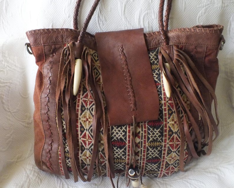 May include: A brown leather tote bag with braided handles and fringe detailing. The bag features a patterned fabric panel with red, black, and beige geometric designs. A leather flap and decorative beads complete the bohemian style.