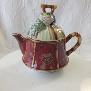 May include: A red and gold teapot with a peacock design on the lid. The teapot has a floral pattern and a bamboo-like handle.