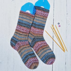 May include: Two pairs of striped socks in shades of brown, blue, and pink, displayed on a white wooden surface with knitting needles and stitch markers.
