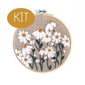 May include: Embroidered daisy flower kit in a wooden hoop. The daisies have white petals and yellow centers, with green stems and leaves. The word "KIT" is in a gold oval.