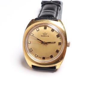 May include: A vintage gold-toned watch with a black leather band. The watch face is gold with black hands and square hour markers. The watch has the name "MARVIN" printed on the face.