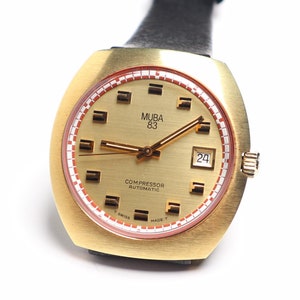 Puede incluir: Un reloj de pulsera dorado con una correa de cuero negro. La esfera del reloj tiene una esfera blanca con marcas negras y el texto "MUBA 63 COMPRESSOR AUTOMATIC" y "24".