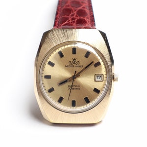 May include: A gold-toned Meister Anker automatic watch with a rectangular face and a date display. The watch features a textured gold dial and a red leather band. The watch face has the text "Automatic 17 Jewels".