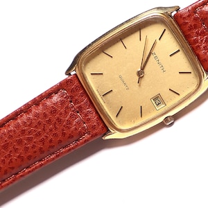 May include: A gold Zenith Quartz watch with a square face and a date display. The watch has a light gold dial and black markers. It is attached to a textured, reddish-brown leather strap. The watch strap is a reddish-brown colour.