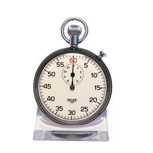 Op de afbeelding: Een vintage Heuer stopwatch met een zwarte behuizing en een witte wijzerplaat. De cijfers zijn zwart, met de 60 in rood. De stopwatch staat op een transparante plastic standaard. Hij meet de tijd in seconden en minuten. De diameter is ongeveer 5 cm.