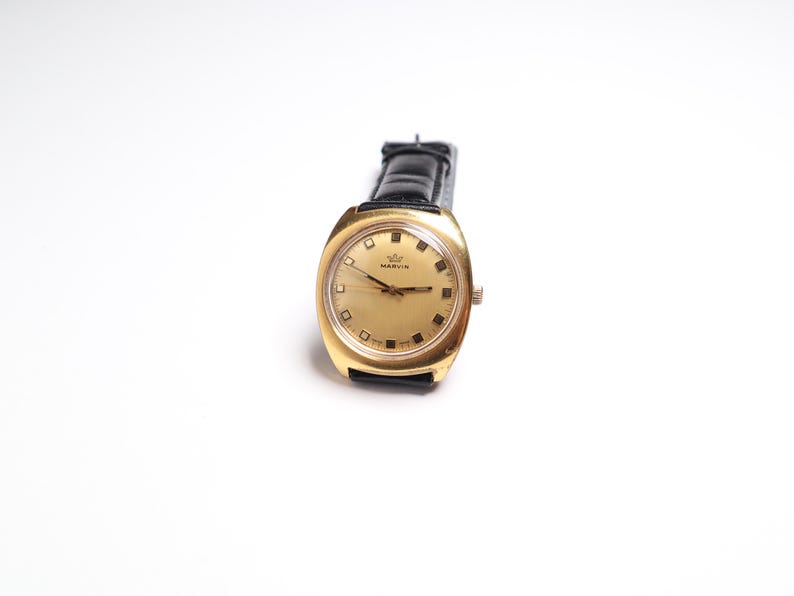 May include: A vintage gold-toned watch with a black leather band. The watch face is gold with black hands and square hour markers. The watch has the name "MARVIN" printed on the face.