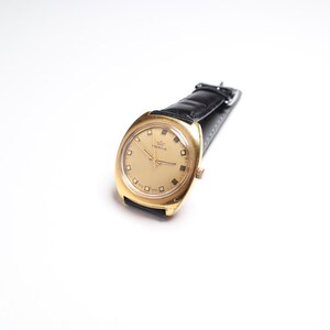 May include: A gold-toned wristwatch with a black leather band. The watch face is a light gold color with square hour markers and a small crown logo. The watch is set against a white background.