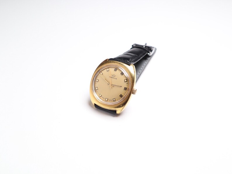 May include: A gold-toned wristwatch with a black leather band. The watch face is a light gold color with square hour markers and a small crown logo. The watch is set against a white background.