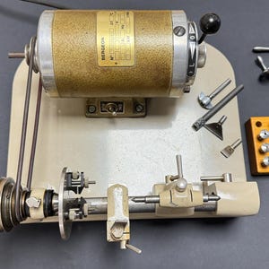 Puede incluir: Un torno de relojero Bergeon con motor dorado, base beige y husillo accionado por correa. El conjunto incluye varios accesorios metálicos, un bloque de madera con veinte pinzas metálicas pequeñas y dos herramientas manuales de metal.