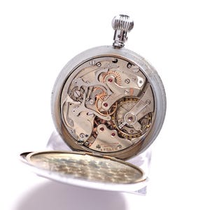 May include: A silver pocket watch with an open face, revealing the intricate mechanical workings of the timepiece. The watch's gears, springs, and other components are visible, showcasing the craftsmanship. The watch is set against a white background.