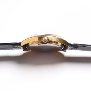 May include: A gold-toned wristwatch with a black leather band. The watch face is round, and the band is secured with a buckle. The watch is viewed from the side, showcasing its profile against a white background.