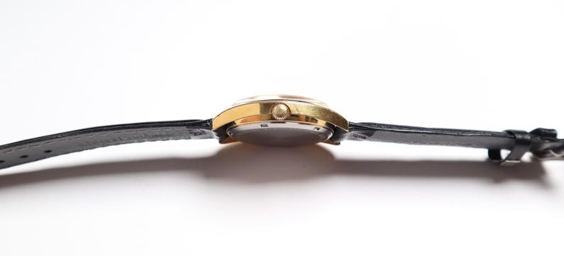 May include: A gold-toned wristwatch with a black leather band. The watch face is round, and the band is secured with a buckle. The watch is viewed from the side, showcasing its profile against a white background.
