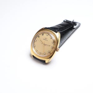 May include: A gold-toned Marvin wristwatch with a black leather band. The watch face is a light gold color with square hour markers and gold hands. The watch is set against a white background.