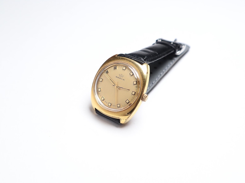 May include: A gold-toned Marvin wristwatch with a black leather band. The watch face is a light gold color with square hour markers and gold hands. The watch is set against a white background.