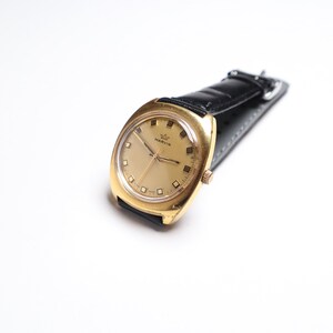 May include: A vintage gold-toned wristwatch with a black leather band. The watch face is gold with rectangular hour markers and the brand name "MARVIN". The watch has a classic, rectangular case design.
