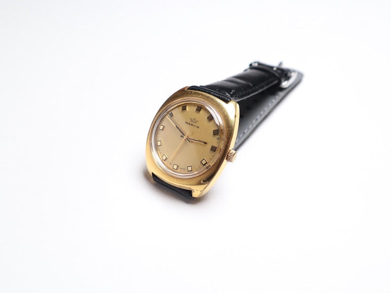 May include: A vintage gold-toned wristwatch with a black leather band. The watch face is gold with rectangular hour markers and the brand name "MARVIN". The watch has a classic, rectangular case design.