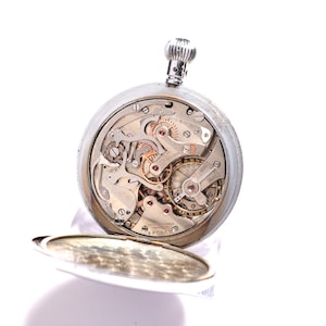 May include: A silver pocket watch with the back open, revealing the intricate mechanical workings of the watch. The watch has a silver winding stem and a hinged cover. The watch's internal gears and springs are visible, showcasing its complex design.