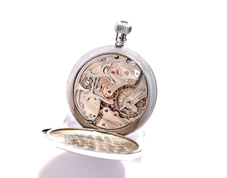 May include: A silver pocket watch with the back open, revealing the intricate mechanical workings of the watch. The watch has a silver winding stem and a hinged cover. The watch's internal gears and springs are visible, showcasing its complex design.