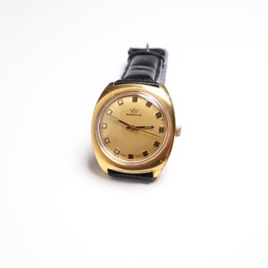 May include: A vintage gold-toned Marvin wristwatch with a black leather band. The watch face is gold with black markers and hands. The watch has a rectangular shape with rounded edges. The word "MARVIN" is printed on the watch face.