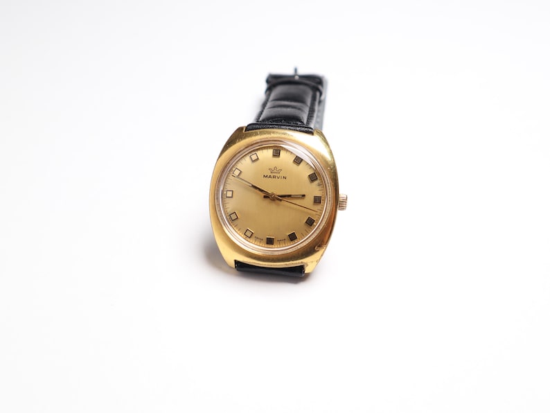 May include: A vintage gold-toned Marvin wristwatch with a black leather band. The watch face is gold with black markers and hands. The watch has a rectangular shape with rounded edges. The word "MARVIN" is printed on the watch face.